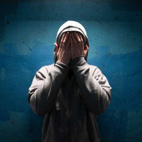 Color image depicting an adult man wearing a grey hoodie with the hood over his head in an underground subway tunnel. His expression is one of despair, sadness and depression. His hands are covering his face in a gesture of bleak desperation and hopelessness. Room for copy space.